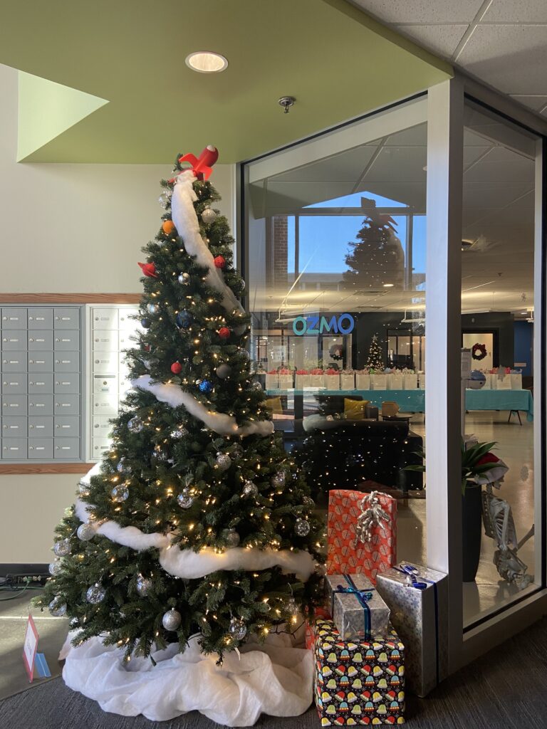 A decorated Christmas tree surrounded by presents in front of an indoor office building titled Ozmo. 